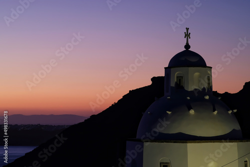 View of a beautiful whitewashed Orthodox Church and Oia in Santorini, while the sun is setting in a dramatic way