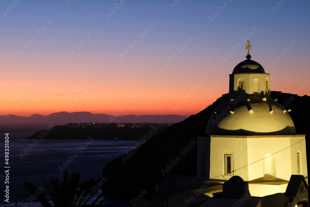 View of a beautiful whitewashed Orthodox Church and Oia Santorini in the background, while the sun is setting in a dramatic way