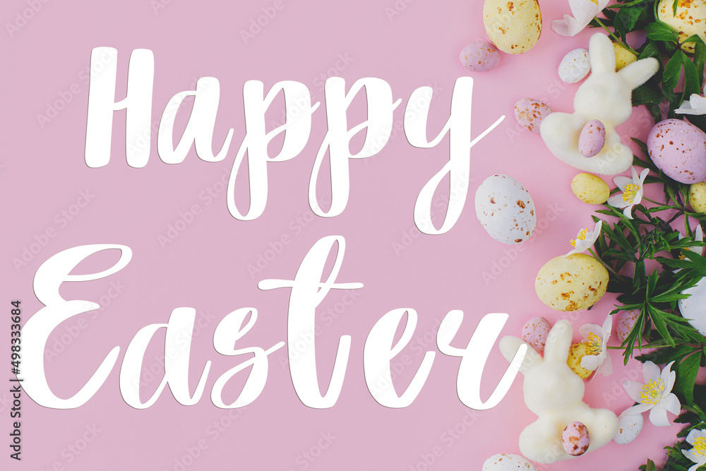 Happy Easter text and Easter chocolate eggs, bunnies and spring flowers border flat lay on pink background. Happy Easter greeting card. Seasons greeting card, handwritten lettering