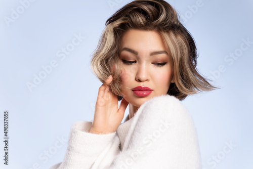 Pretty stylish young asian woman with stylish short haircut wearing cozy white sweater posing on blue studio background. Haircare, hairstyling cosmetics advertisement. Natural beauty.