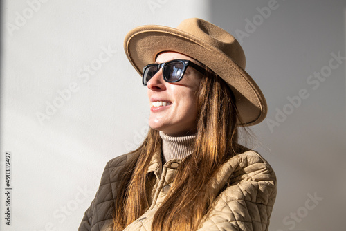 Smiling young woman in a jacket in a hat in sunglasses against a white wall.