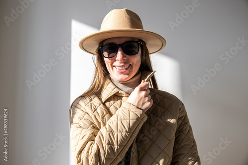 Smiling young woman in a jacket in a hat in sunglasses against a white wall.