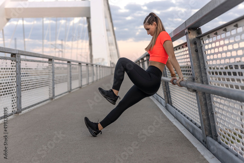 Young happy focused fitness girl in black yoga pants and orange short shirt work out and stretch her body on the bridge footpath during the day. Front view.