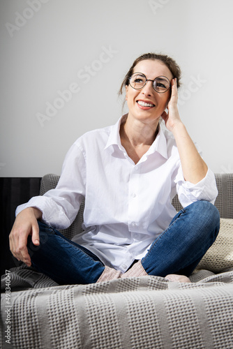 Smiling young woman in glasses in a white shirt sitting on a sofa against the background of white walls.