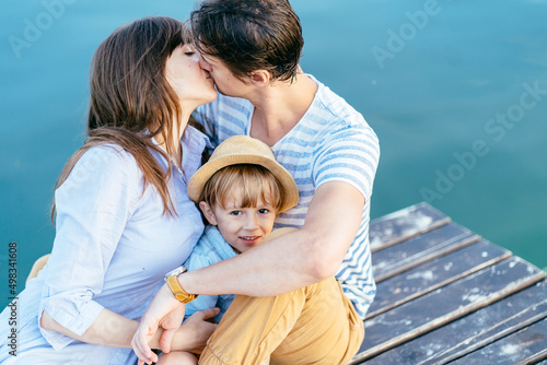 Mother and father kissing while their little son on wooden bridge over blue water on background. Happy vacation holiday concept. © Iryna