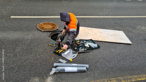 A road worker sits on the edge of a manhole with tools and materials spread out around.