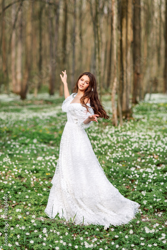 girl in a white lace dress dances in a clearing with primroses. A young woman with long hair in a spring forest.