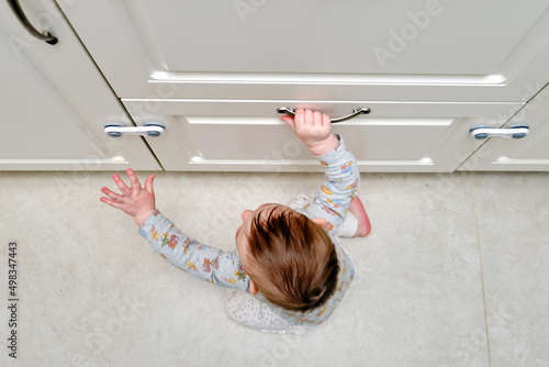 Toddler baby boy rips off a cabinet drawer with his hand. The child holds the cabinet door handle, small kid photo