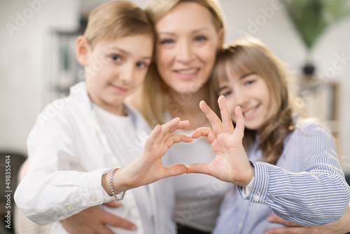 Cute little daughter with son and mother join hands in shape of heart as concept of mom and kids love care support, smiling mum and her kids looking at camera posing together for headshot portrait