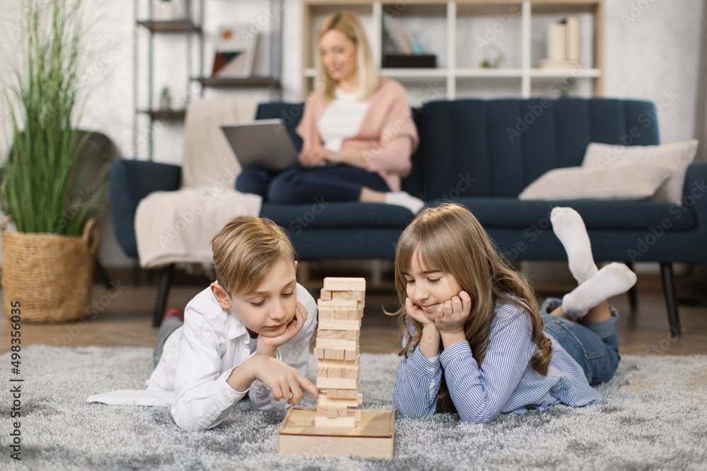 Little children sit on warm floor in living room play construct with wooden building blocks bricks together make a tower, young mom rest on sofa in background, Caucasian family relax at home Little children sit on warm floor in living room play construct with wooden building blocks bricks together make a tower, young mom rest on sofa in background, Caucasian family relax at home