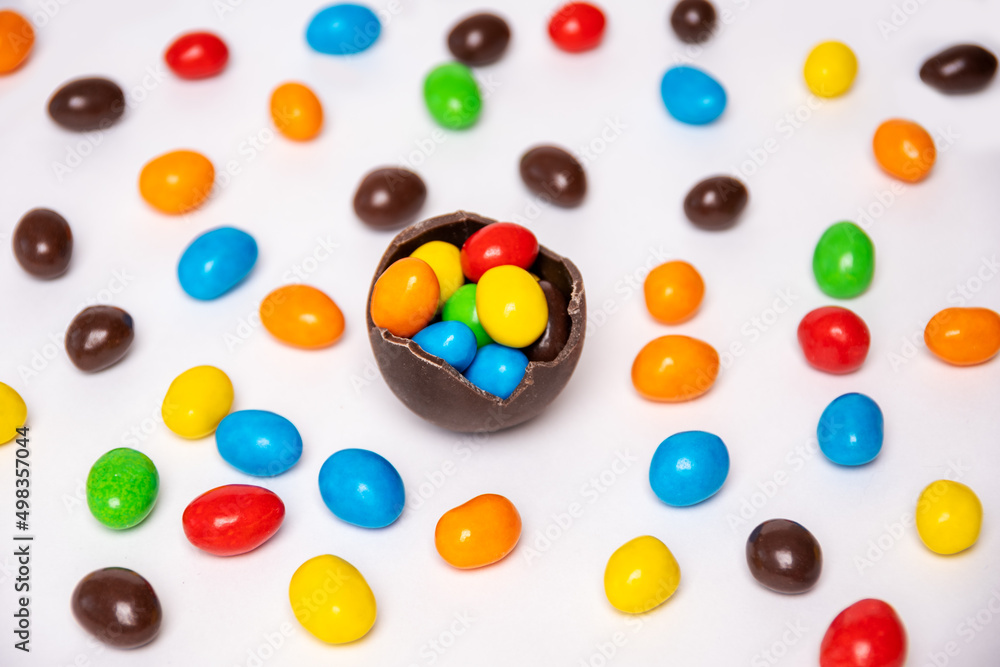 Half of a cracked chocolate egg filled with multi-colored candies on a white background with multi-colored candies, top view. Treats for children. Sweet dessert for kids. Sweet Easter tradition. Half of a cracked chocolate egg filled with multi-colored candies on a white background with multi-colored candies, top view. Treats for children. Sweet dessert for kids. Sweet Easter tradition.
