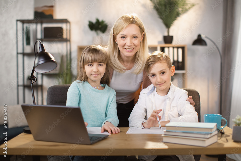 Portrait of joyful small daughter, son and their mother doing homework and looking at camera. Happy young mom praising little preschool children girl and boy for making right tasks, sitting at home. Portrait of joyful small daughter, son and their mother doing homework and looking at camera. Happy young mom praising little preschool children girl and boy for making right tasks, sitting at home.