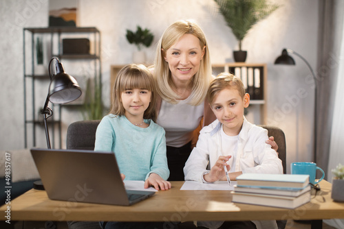 Portrait of joyful small daughter, son and their mother doing homework and looking at camera. Happy young mom praising little preschool children girl and boy for making right tasks, sitting at home.