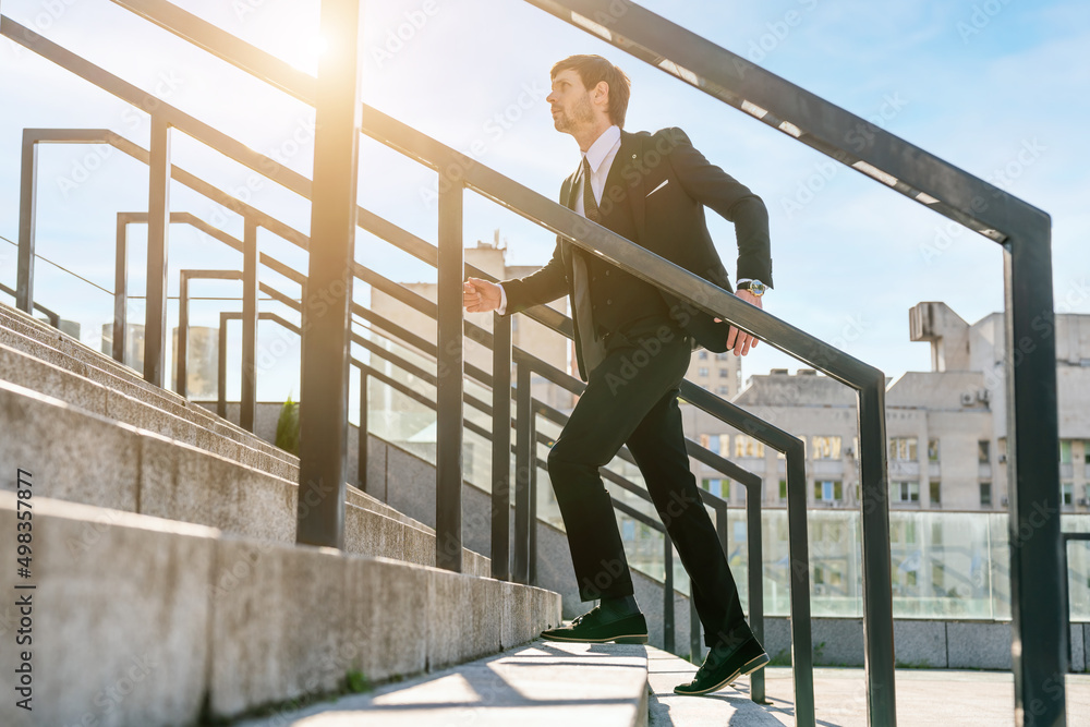 Portrait of a young and handsome businessman wearing suit jacket running fast up the stairs. Stylish successful man go to work in the morning city with office building in the background. Side view. Portrait of a young and handsome businessman wearing suit jacket running fast up the stairs. Stylish successful man go to work in the morning city with office building in the background. Side view.