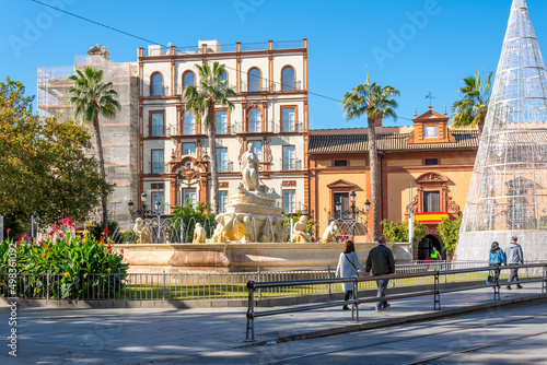 Unidentifiable edestrians pass by the Fountain of Seville in the Puerta de Jerez square in the historic center of Seville, Spain. 