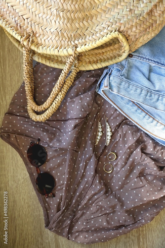 Brown polka dot blouse, vintage mom jeans, straw bag, round sunglasses, gold rings and pearl hair clips. Fashionable outfit on wooden background. Flat lay. 