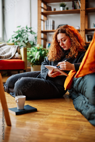 A young ginger woman is writing in her notebook while sitting on the floor in the living room and having a cup of coffee.