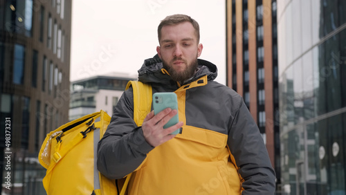 Portrait of restaurant food delivery guy delivering an order. Courier searches for delivery address on street. Handsome man with smartphone delivers food in yellow thermos bag in open air.
