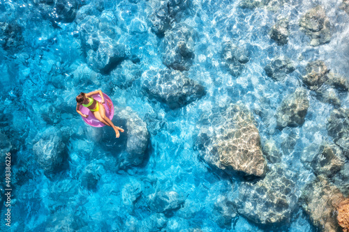 Aerial view of a young woman swimming with pink swim ring in blue sea at sunset in summer. Tropical landscape with girl, clear water, stones, sandy beach. Top view. Vacation in Lefkada island, Greece