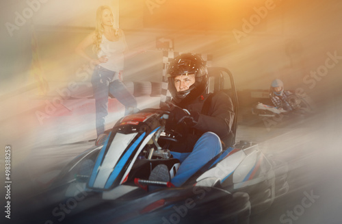 Diligent efficient positive man in helmet driving car for karting in sport club, woman with flag on background © JackF