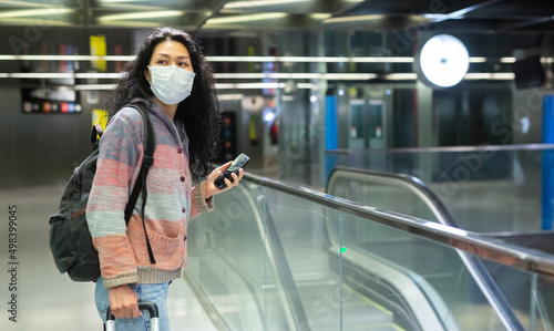 Portrait of positive female traveler in protective face mask walking in hall of metro station