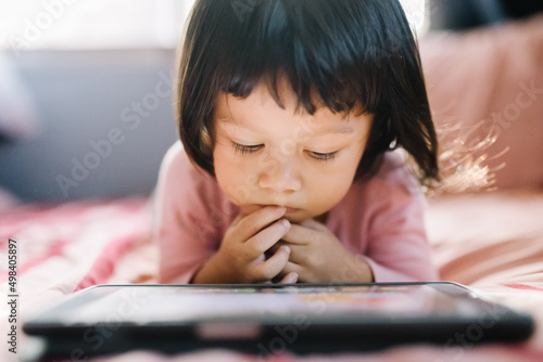 A little girl in a pink shirt lay intently playing the tablet on her bed. self-learning technology education concept