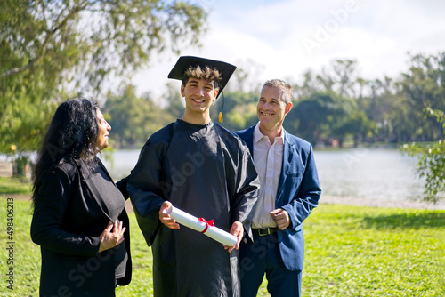 Young recently graduated boy, dressed in cap and gown, with his degree in hands, celebrating with his multi ethnic  family on the university campus. Very happy expression, achievement photo