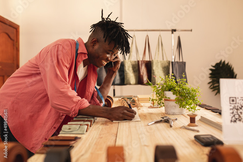 Smiling young african american male owner writing on book during phone call in leather store photo