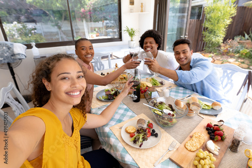 Smiling young biracial woman taking selfie while toasting drinks at dining table photo