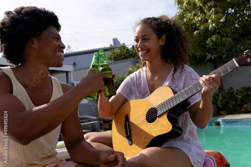 Happy biracial woman with guitar toasting beer bottle with man at poolside on sunny day photo