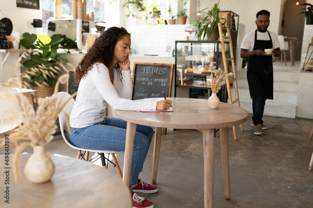 African american female customer writing in diary while barista with coffee in background at cafe African american female customer writing in diary while barista with coffee in background at cafe