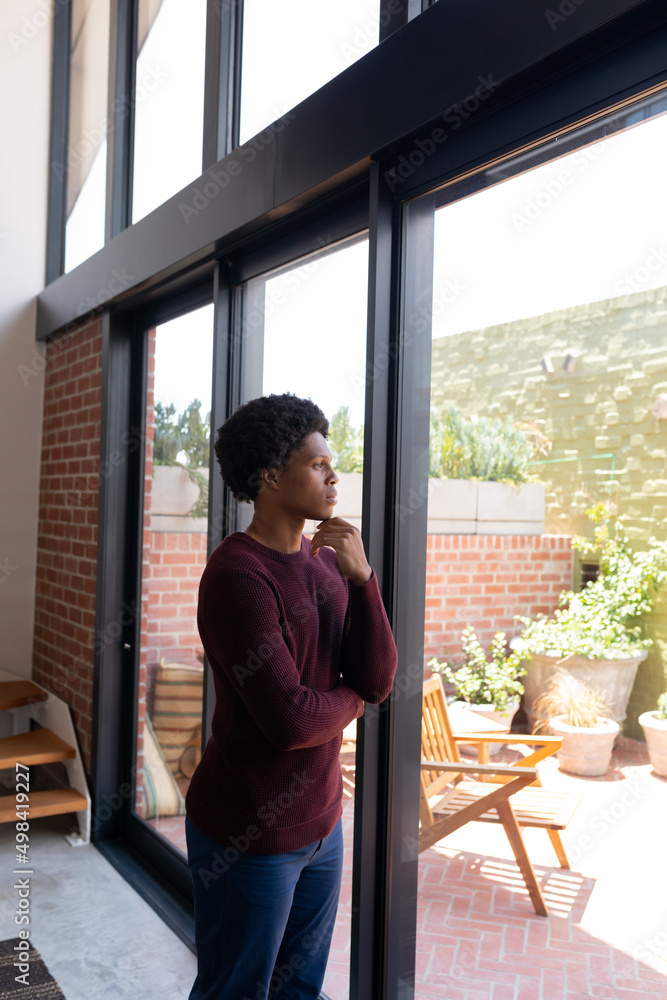 African american young man with hand on chin looking through window while standing at home African american young man with hand on chin looking through window while standing at home