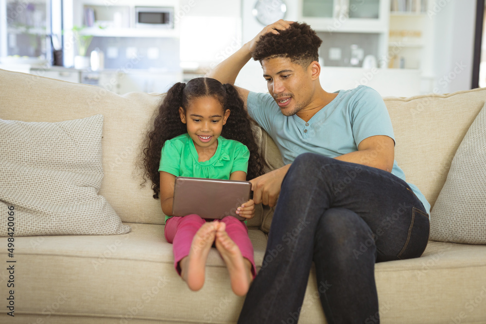 Biracial man helping daughter in digital tablet while sitting together on sofa at home Biracial man helping daughter in digital tablet while sitting together on sofa at home