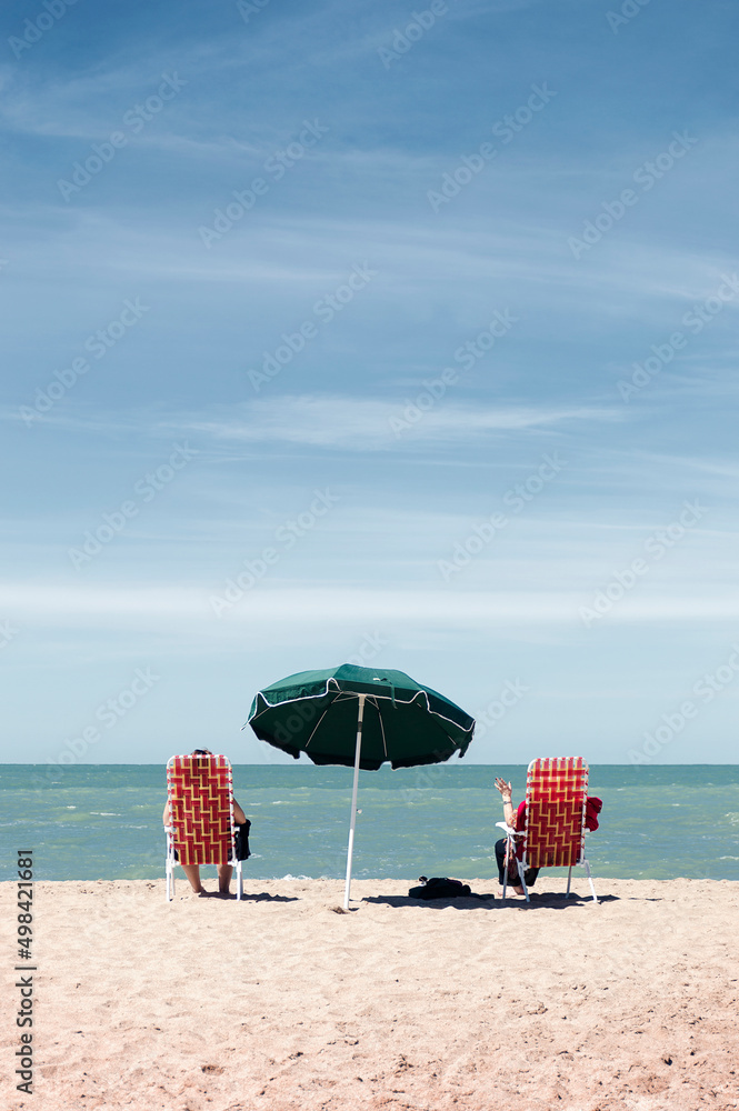 Two women sitting on deck chairs under an umbrella facing the sea