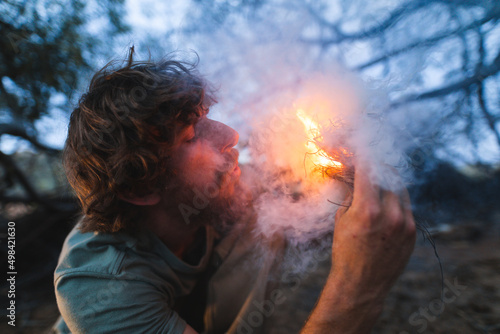 Young male caucasian survivalist blowing on fire and smoke in forest photo