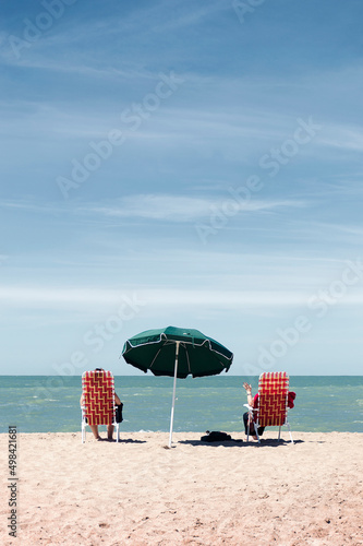 Two women sitting on deck chairs under an umbrella facing the sea