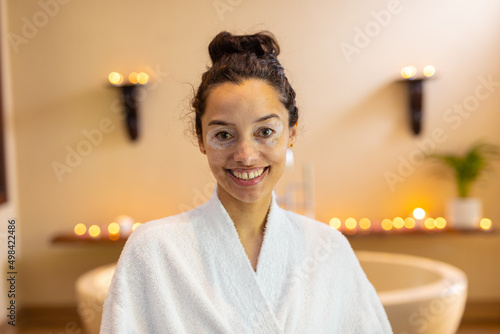 Portrait of smiling biracial young woman wearing bathrobe standing in front of bathtub at spa photo