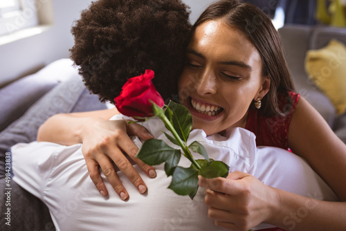 Happy young biracial woman holding red rose hugging boyfriend at home photo