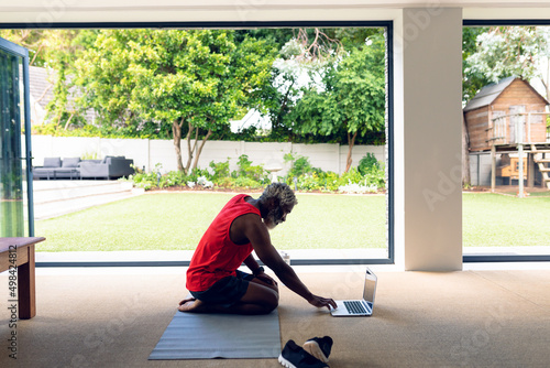 African american man using laptop while kneeling on yoga mat in living room at home photo