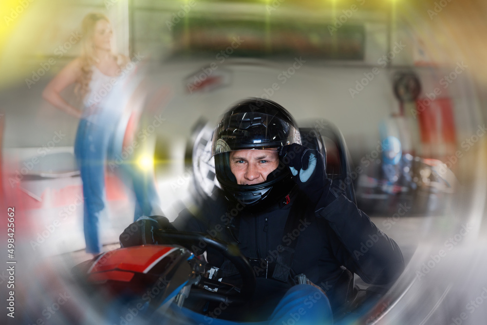 Diligent efficient positive man in helmet driving car for karting in sport club, friendly smiling woman with flag on background