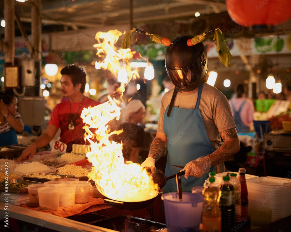 Hot stuff. Shot of an unidentifiable food vendor wearing a helmet while flambeing something in a pan.