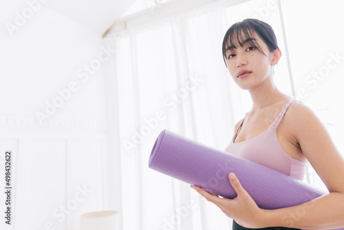 Smiling Asian woman wearing sportswear and yoga pants holding a rolled-up yoga mat standing in a bedroom with sunlight from the window. Image with copy space.