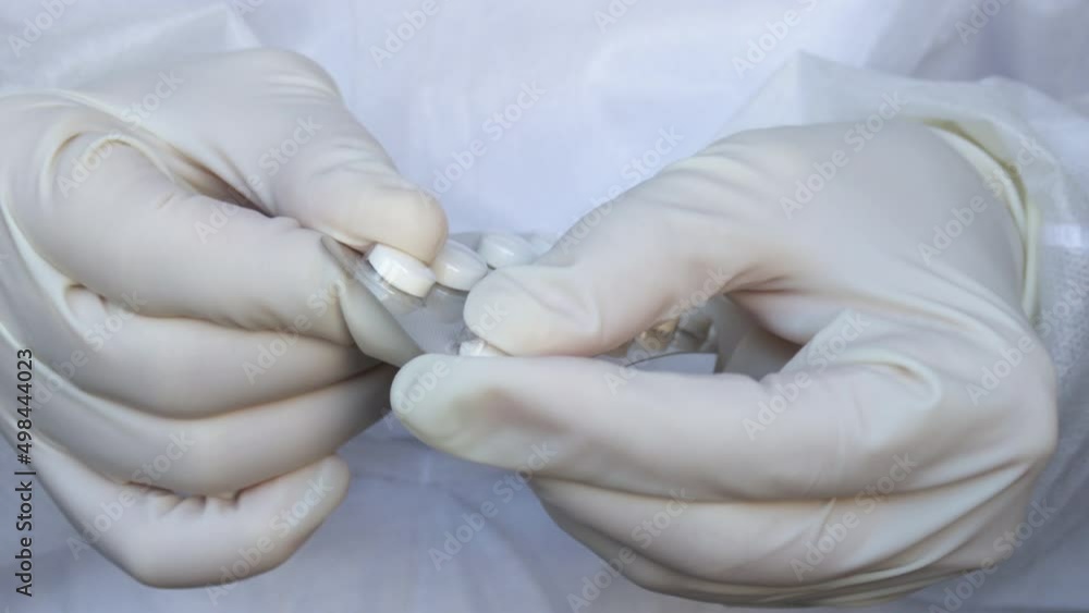 woman's (doctor, nurse) hands in gloves taking out pill from blister pack. medical assistant in white disposable robe is holding plastic blister with round tablets. Natural herbs, organic pills woman's (doctor, nurse) hands in gloves taking out pill from blister pack. medical assistant in white disposable robe is holding plastic blister with round tablets. Natural herbs, organic pills