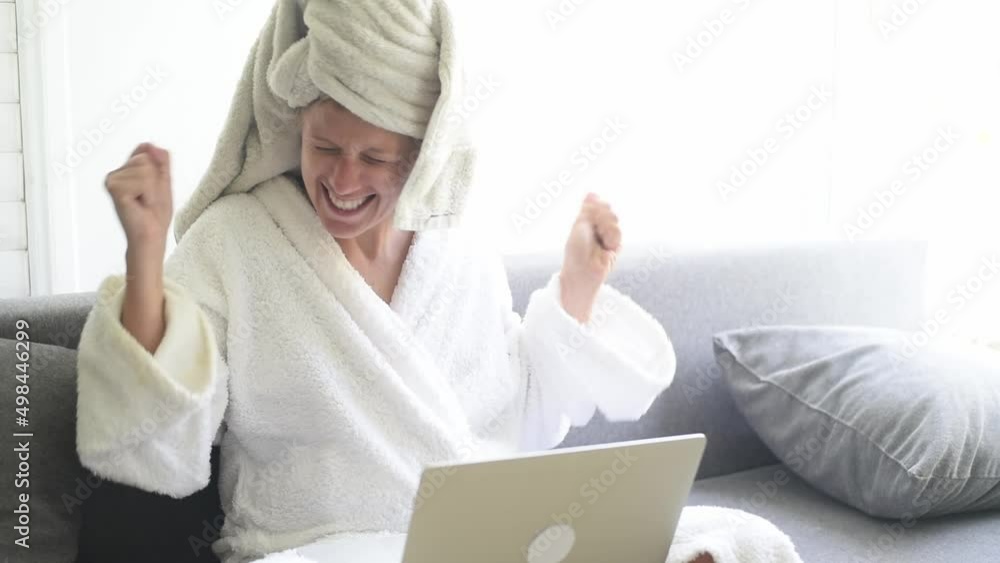 Young woman in white robe and with towel on head sits on couch and work on laptop. Person talk online, chatting with colleagues and joys of success. Girl work on home Young woman in white robe and with towel on head sits on couch and work on laptop. Person talk online, chatting with colleagues and joys of success. Girl work on home