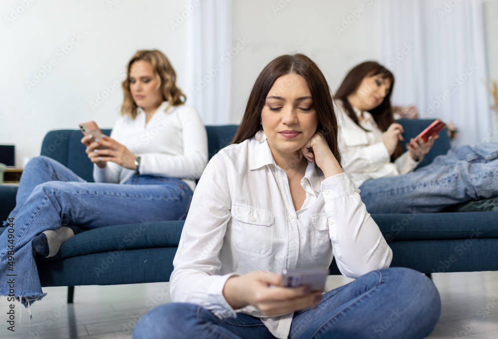 Three girls - friends in shirts and jeans sit on and near the sofa in the room and climb social networks