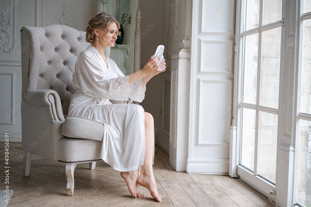Attractive caucasian woman touching healthy flawless radiant smooth face skincare while looking in mirror while sitting in armchair in living room wearing white bathrobe, skincare and makeup concept
