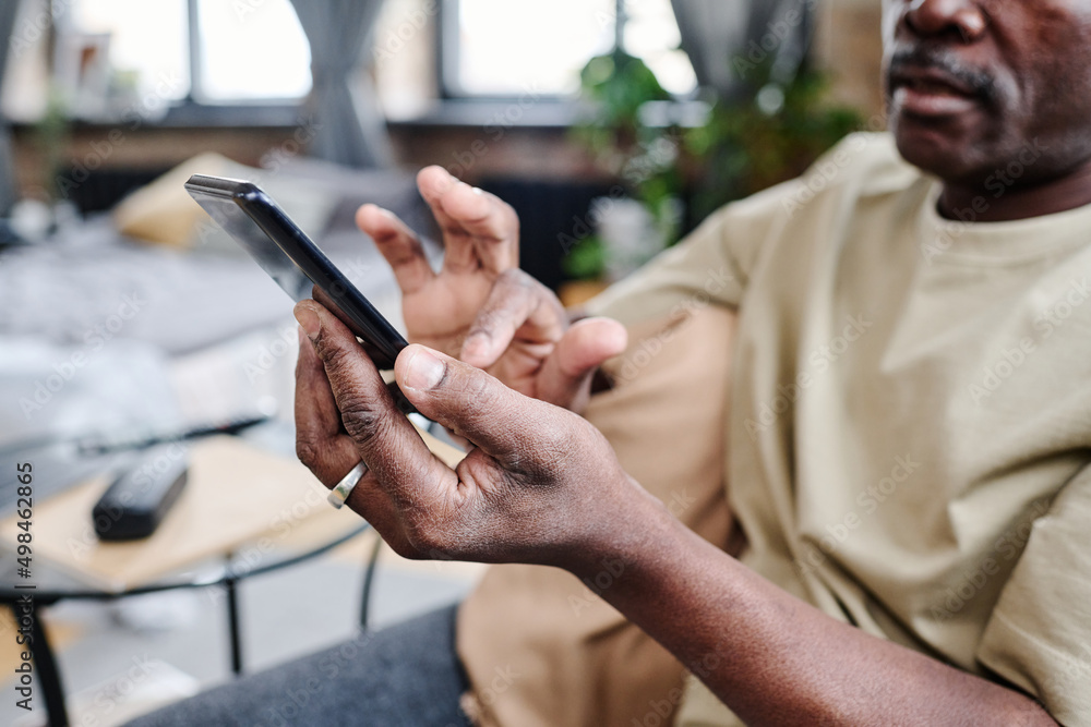 Obraz premium Hands of senior African American man in home wear texting in mobile phone while relaxing in living room of loft apartment