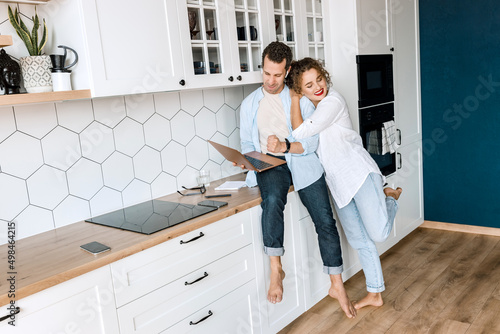 Lovely family couple are standing at the kitchen at home, doing online shopping or chatting with friends. Joyful stylish husband and wife using laptop, watching funny videos or photos, smiling