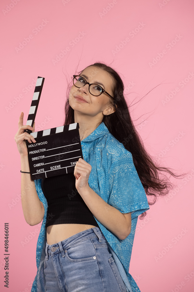 Young attractive woman holding film clapper. Teen girl in a blue shirt and glasses holding classic black film making clapperboar on a pink background. Young attractive woman holding film clapper. Teen girl in a blue shirt and glasses holding classic black film making clapperboar on a pink background.