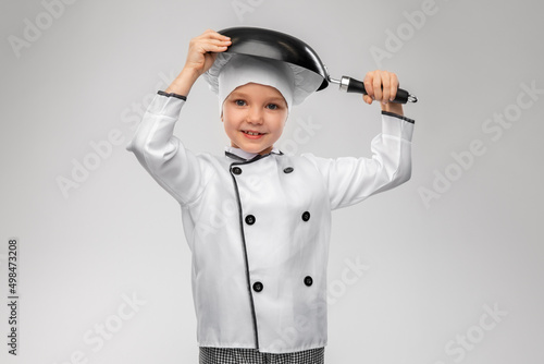 cooking, culinary and profession concept - happy smiling little girl in chef's toque and jacket with frying pan over grey background